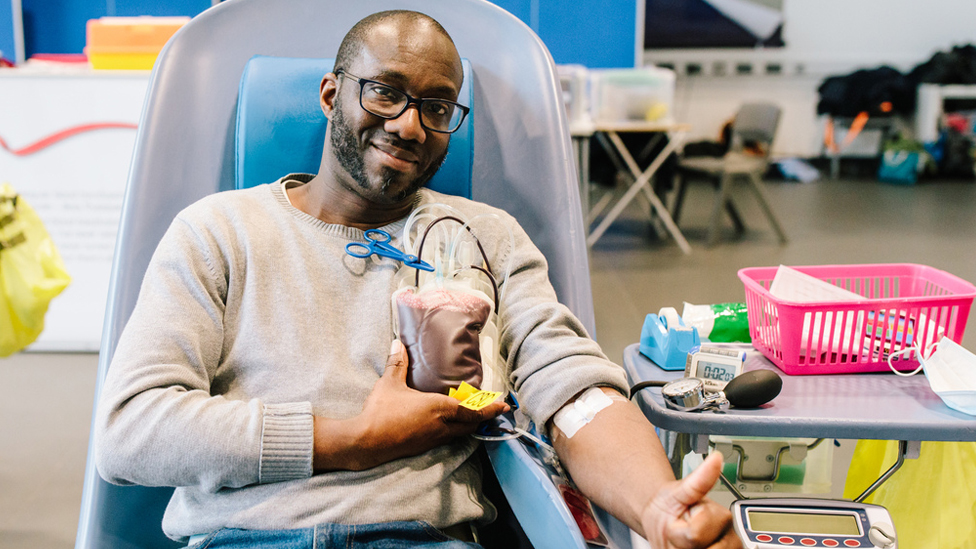 A man with glasses smiles and gives a thumbs up while donating blood, with a collection bag visible. He is seated in a blood donation chair in a well-lit environment.
